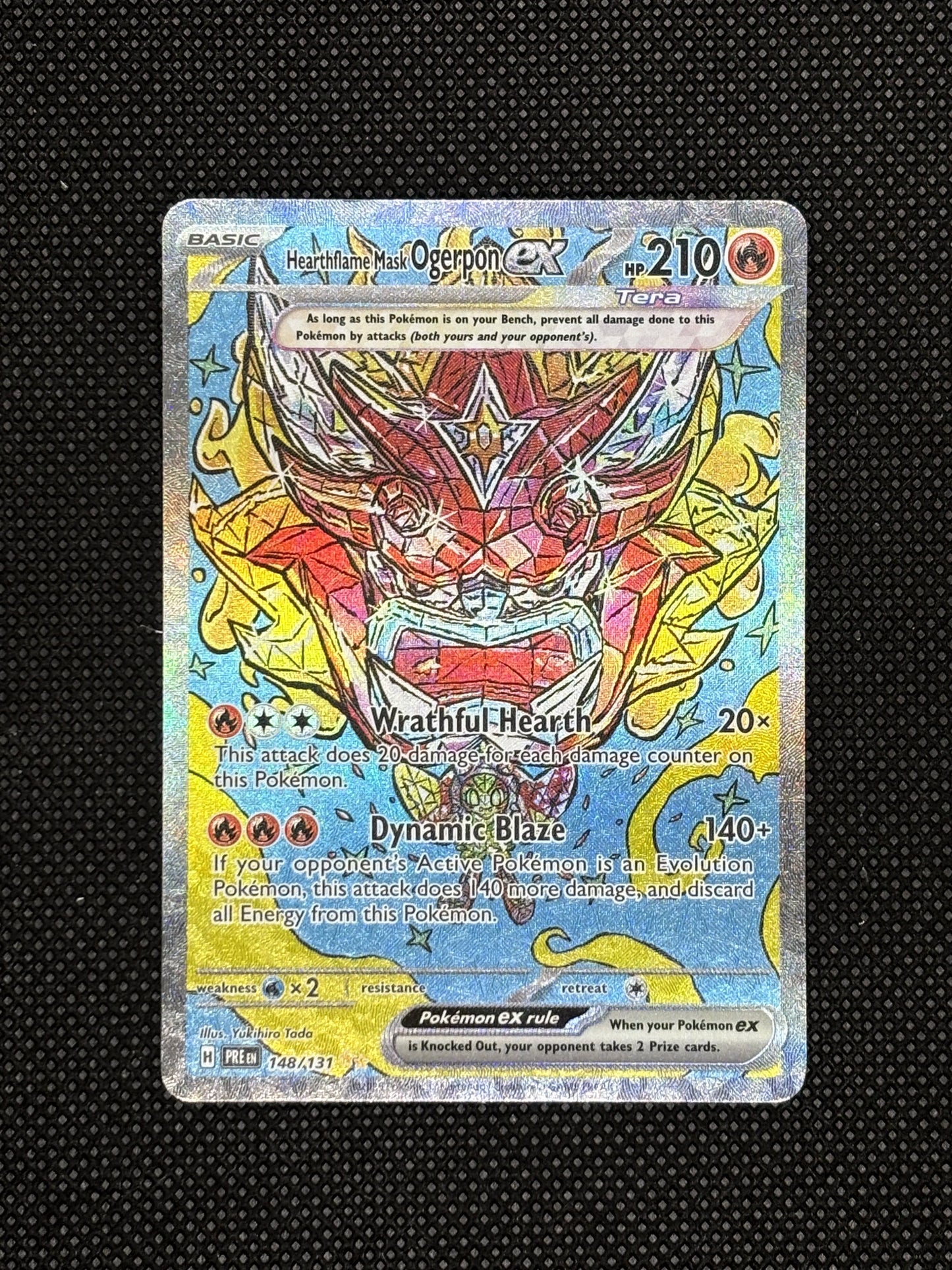 Hearthflame Mask Ogerpon EX from Prismatic Evolutions with a red mask, yellow and blue background, and is textured.