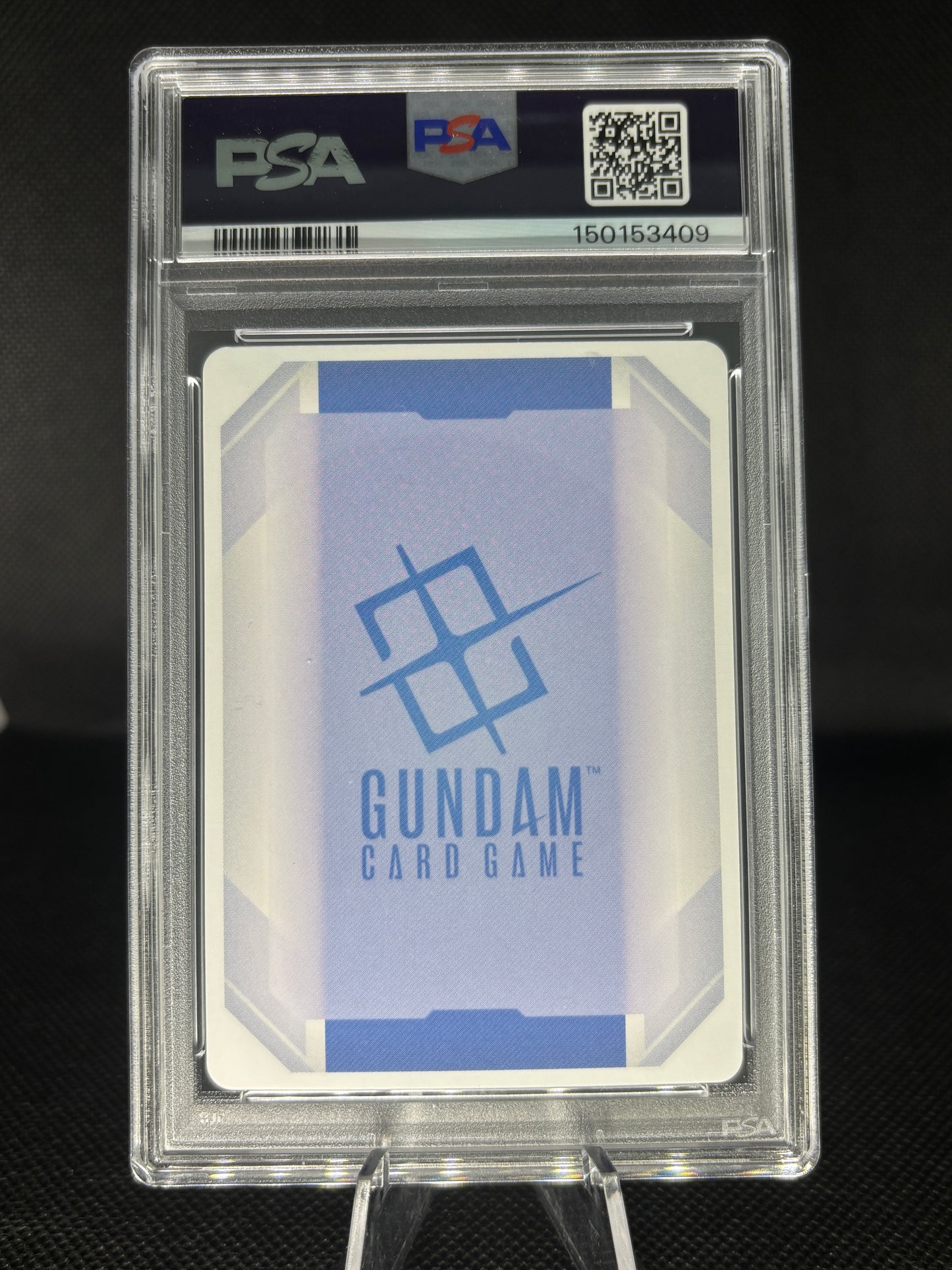 A PSA graded Gundam Card Game card in a clear PSA slab, displayed on a stand against a dark background. The card features a blue and white geometric design with the Gundam logo and text.