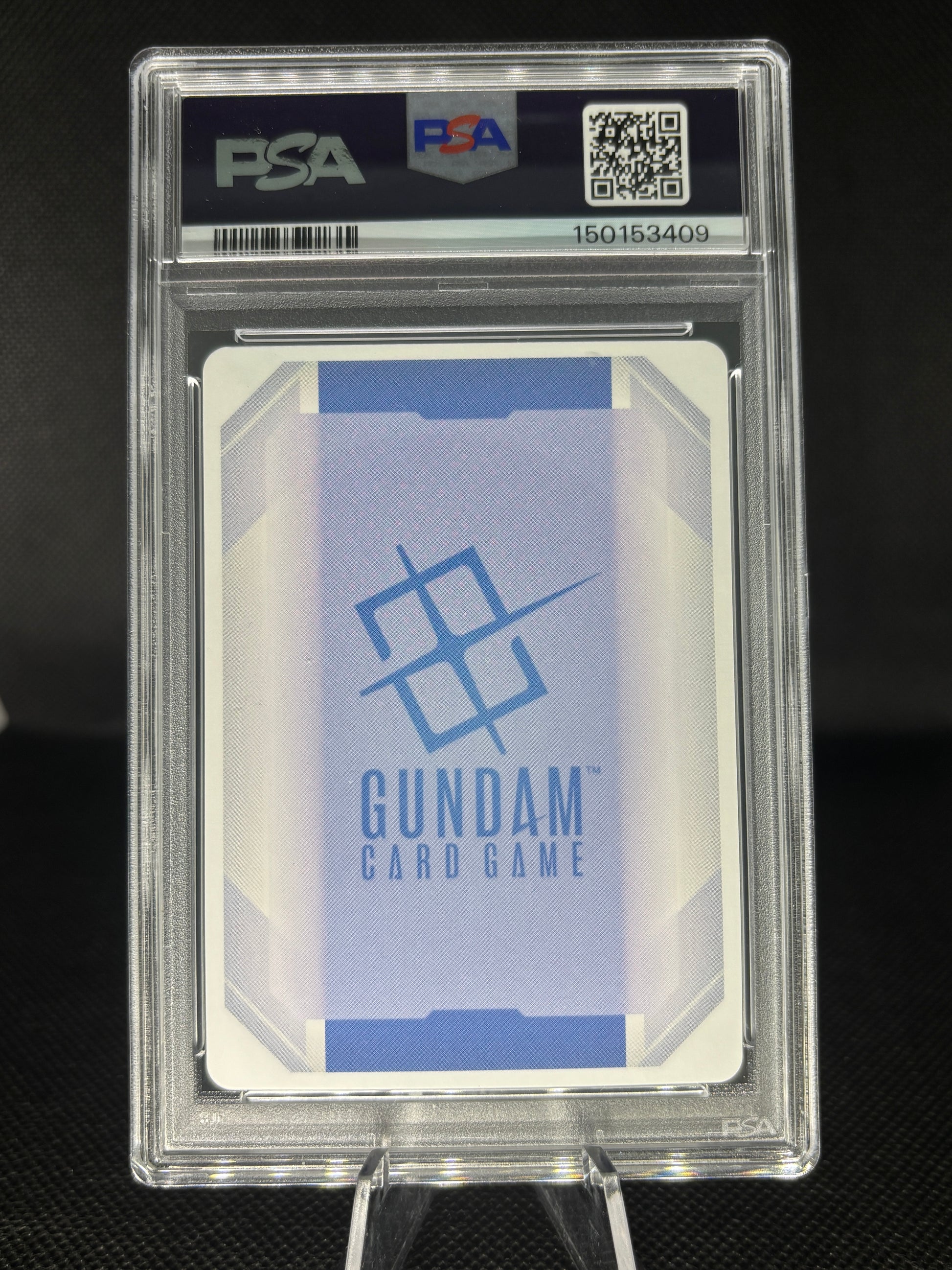 A PSA graded Gundam Card Game card in a clear PSA slab, displayed on a stand against a dark background. The card features a blue and white geometric design with the Gundam logo and text.